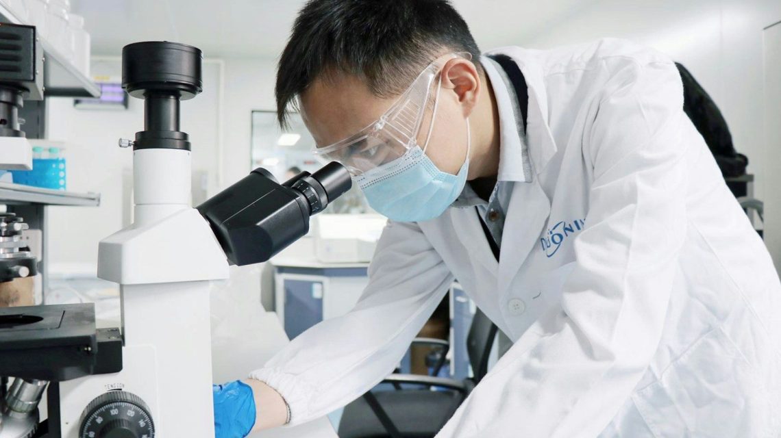 A man in a lab coat and goggles looking through a microscope