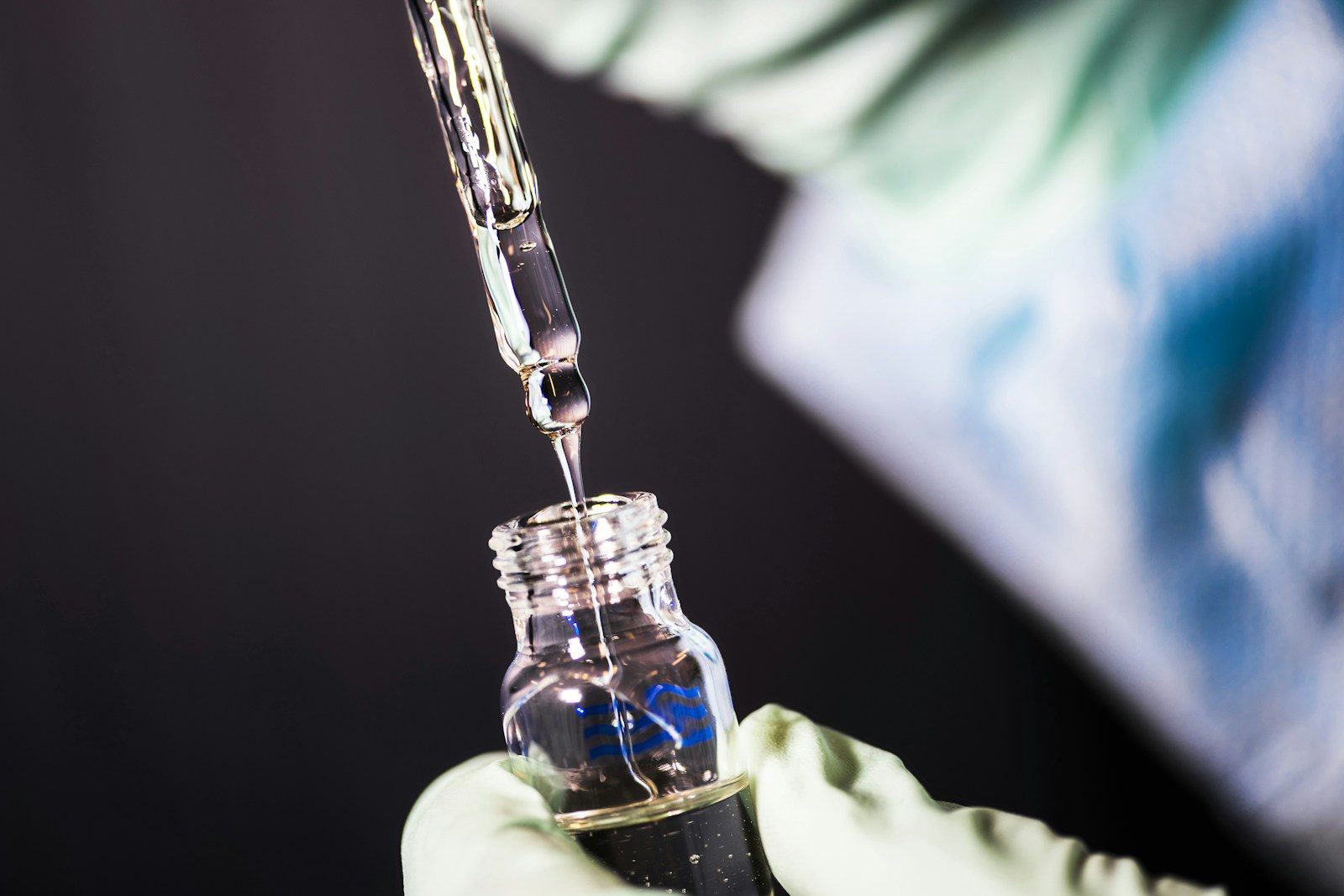 A person in green gloves dripping liquid from a glass pipette into a vial